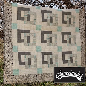 May include: A white quilt with a geometric pattern of gray squares and aqua blue accents. The quilt is hanging on a clothesline in front of a tree. The quilt is labeled "Sweetwater".