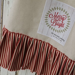 May include: A cream-colored fabric Christmas stocking with a red and white striped ruffle at the bottom. The stocking has a rectangular patch with the text "Merry and Bright" in red cursive lettering with a green wreath around it. The text "the burnett family" is below the wreath in black lettering.