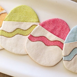 Puede incluir: Cuatro posavasos de tela con forma de huevo de Pascua, con base blanca y parte superior de colores. Los posavasos tienen un diseño de puntadas marrones onduladas alrededor del borde.