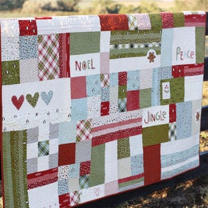 Pode incluir: Uma colcha de retalhos com uma variedade de blocos de tecido retangulares em vermelho, verde, branco e azul claro. A colcha inclui texto impresso lendo NOEL, PEACE e JINGLE, juntamente com motivos de corações bordados e pequenos padrões de pinguins.