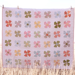 May include: A handmade quilt with a repeating floral pattern in pastel colors. The flowers are arranged in a grid, with each flower composed of eight fabric pieces. The quilt is hanging on a clothesline in a field of tall grass.