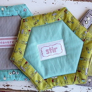 Könnte beinhalten: Drei sechseckige Topflappen aus Stoff mit gestreiftem Hintergrund und buntem Rand. Die Topflappen sind mit den Bezeichnungen "messen", "rühren" und "mischen" versehen.