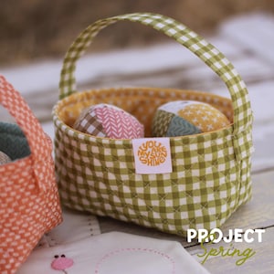 May include: A small, quilted basket with a green and white checkered pattern, holding two decorative fabric eggs. The basket has a handle and a yellow interior. A tag on the basket reads "YOU ARE MY SUN SHINE".