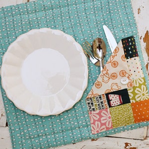 May include: A white scalloped plate rests on a teal placemat with a white geometric pattern. Silverware sits next to a patchwork utensil holder with various fabric patterns, including bicycles and floral designs.