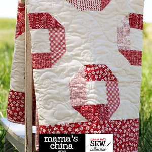 May include: A red and white quilt with a patchwork design. The quilt is made of various fabrics, including gingham, floral prints, and solid red. The quilt is hanging on a wooden chair. The quilt is labeled "mama's china" and "Sweetwater".
