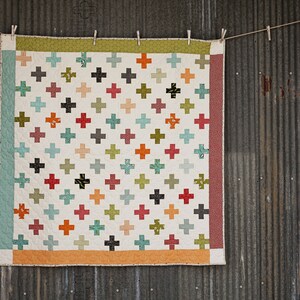 May include: A white quilt with a patchwork design featuring a variety of colourful plus signs. The quilt is hanging on a rustic metal wall.