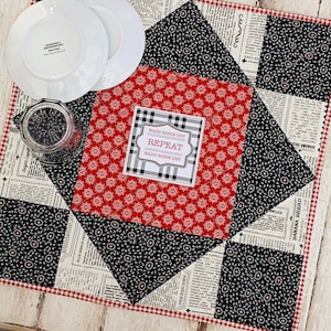 May include: A red and black quilted table runner with a white gingham border. The runner has a black and white newspaper print fabric and a red fabric with a white floral pattern. A white tag with black text reads "Wash Rinse Dry Repeat Wash Rinse Dry".