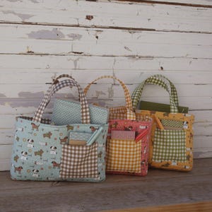 Pode incluir: Três sacolas de tecido com bolsas combinando. As sacolas apresentam padrões de cães e xadrez vichy em azul, rosa e amarelo. Cada sacola tem um bolso e alças. As sacolas estão em uma superfície de madeira.