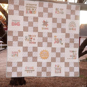 May include: A handmade quilt with a checkerboard pattern in cream and brown. The quilt features embroidered inspirational phrases like "Dream Big," "Shine," and "Choose Joy." The quilt is hanging outdoors, showcasing its homespun charm.