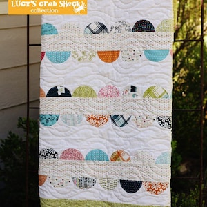 May include: A white beach blanket quilt with a patchwork design of colorful half-circles. The quilt is hanging on a metal fence and has the text "beach blanket #P189 LUCY'S crab SHACK collection Sweetwater" on it.