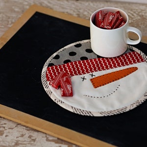May include: A white ceramic mug filled with red sweets, resting on a snowman-shaped fabric coaster with black and white polka dots and red and white stripes. The coaster is placed on a black chalkboard surface.