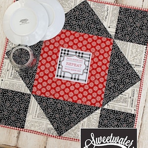 May include: A red and black quilted table runner with a white and black checkered border. The runner features a newspaper print fabric with a small red and white floral pattern. A white card with black text reads "WASH RINSE DRY REPEAT WASH RINSE DRY". The text "Sweetwater" is written in white on a black background.