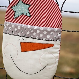 Könnte beinhalten: Ein roter und weiß gestreifter Schneemann-Topflappen mit einem grau-weißen gepunkteten Hut, einer orangefarbenen Filznase und einem schwarzen gestickten Lächeln.