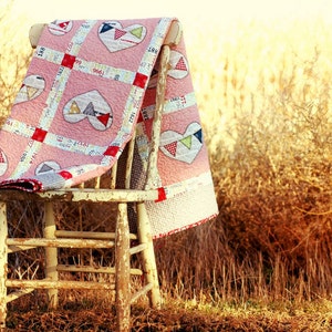 May include: A pink and red patchwork quilt with hearts and flags draped over a weathered wooden chair. The chair is sitting in a field of tall, dry grass.