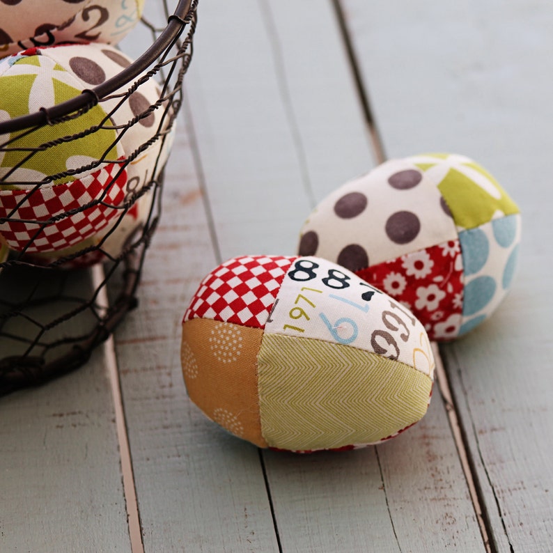 May include: Three patchwork fabric Easter eggs, two are on a light blue wooden surface and one is in a wire basket. The eggs are made of different colored fabrics with geometric patterns and numbers.