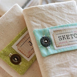 May include: Three small, cream-colored canvas notebooks with decorative labels. The labels are green, blue, and white with a floral pattern and the words "jot" and "sketch".