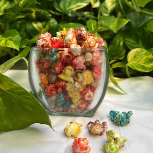 May include: A glass bowl filled with colorful popcorn. The popcorn is a mix of red, yellow, blue, green, and white. There are also a few pieces of popcorn scattered on a white surface.