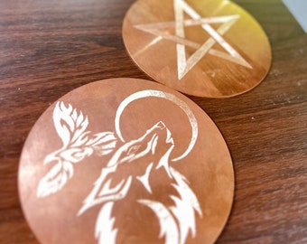 May include: Two copper coasters with etched designs. One coaster features a wolf howling at the moon, the other features a pentagram.