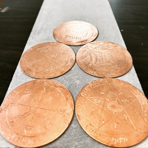 Puede incluir: Cinco posavasos redondos de cobre con intrincados diseños y símbolos grabados en la superficie. Los posavasos están dispuestos en semicírculo sobre una superficie gris claro.