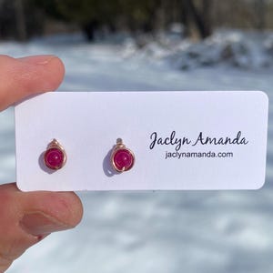 May include: A pair of earrings with small, round, pink gemstones wrapped in gold wire. The earrings are displayed on a white card with the text "Jaclyn Amanda" and "jaclynamanda.com".