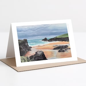 Puede incluir: Una tarjeta de felicitación con una pintura de paisaje costero. La obra de arte representa una playa con acantilados rocosos, una orilla arenosa y olas del océano bajo un cielo nublado. La tarjeta se muestra sobre una superficie blanca con un sobre marrón.