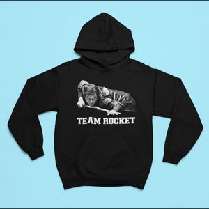Puede incluir: Sudadera con capucha negra con un gráfico blanco de un hombre jugando al billar y el texto "TEAM ROCKET".