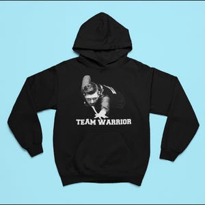 Puede incluir: Sudadera con capucha negra con un gráfico blanco de una persona jugando al billar. El gráfico incluye el texto "TEAM WARRIOR".