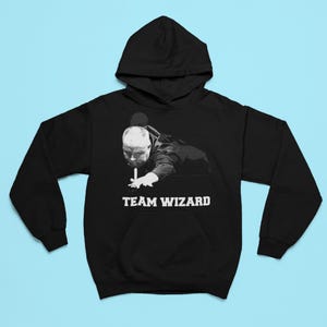 Puede incluir: Sudadera con capucha negra con un gráfico blanco de un hombre jugando al billar. El gráfico incluye el texto "TEAM WIZARD".