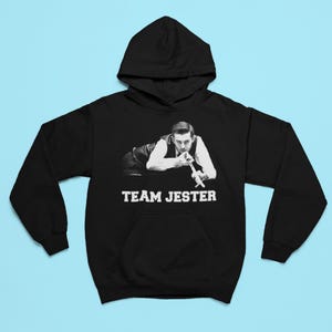 Puede incluir: Sudadera con capucha negra con bolsillo delantero y capucha. El gráfico frontal presenta una imagen en blanco y negro de un hombre sosteniendo un taco de billar, con el texto "TEAM JESTER" en blanco.