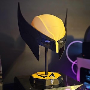 Puede incluir: Una máscara de Wolverine amarilla y negra con grandes aletas laterales puntiagudas y una franja central amarilla, montada sobre un soporte vertical negro con una base hexagonal amarilla que muestra un logo negro y tres marcas de garras blancas.