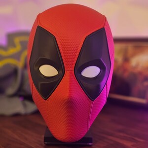 May include: A red and black Deadpool mask with white eyes. The mask is on a black stand.