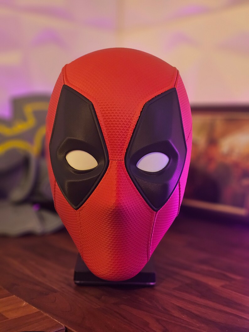 May include: A red and black Deadpool mask with white eyes. The mask is on a black stand.