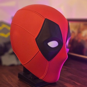 May include: A red and black Deadpool mask with a white eye detail. The mask is on a black stand.
