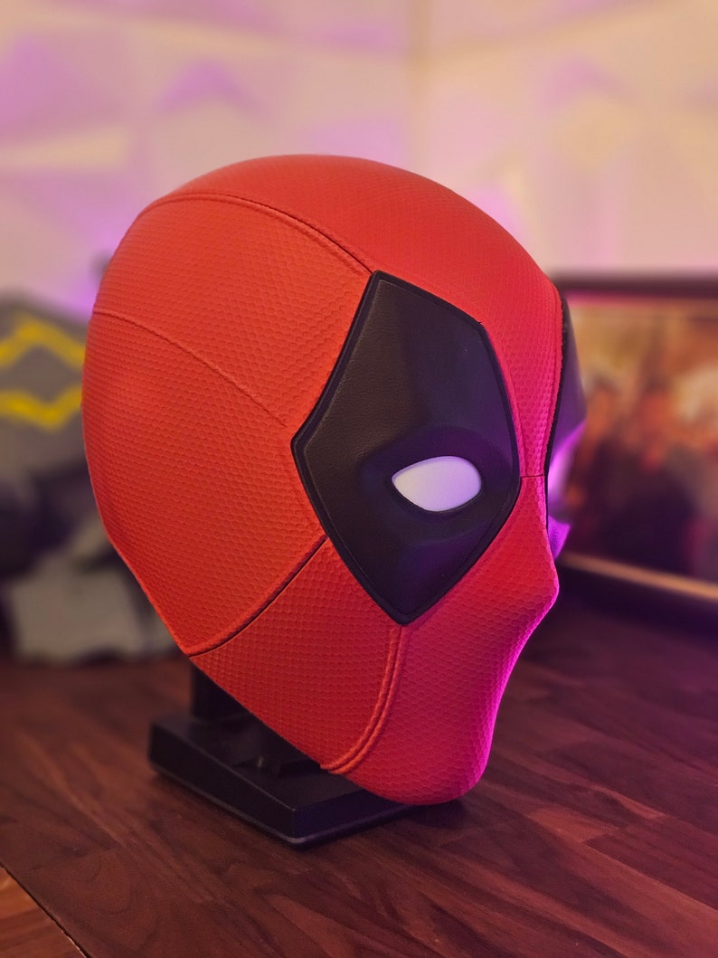May include: A red and black Deadpool mask with a white eye detail. The mask is on a black stand.