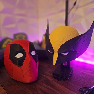 May include: A red and black Deadpool mask and a yellow and black Wolverine mask. The masks are on a wooden surface with purple lighting.