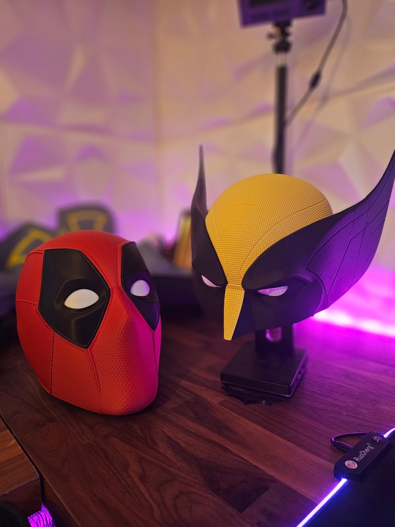 May include: A red and black Deadpool mask and a yellow and black Wolverine mask. The masks are on a wooden surface with purple lighting.