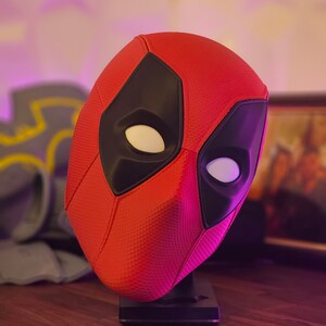 May include: A red and black Deadpool mask on a black stand. The mask has a textured surface and white eye holes.