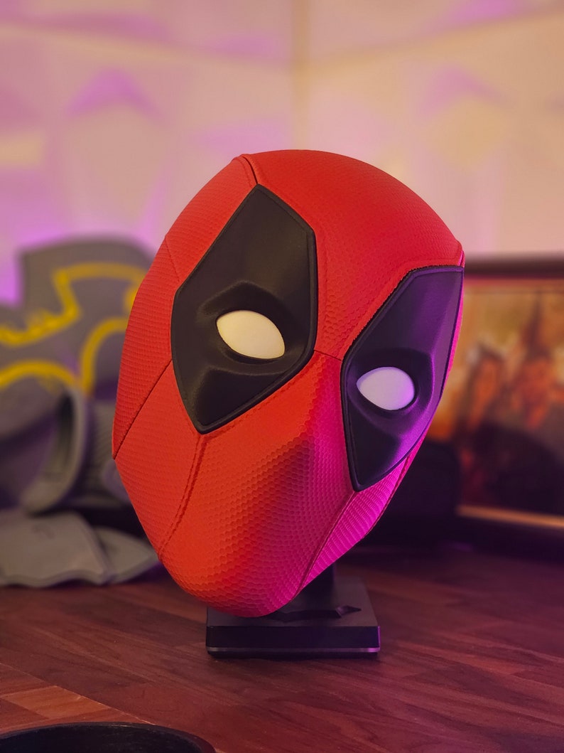 May include: A red and black Deadpool mask on a black stand. The mask has a textured surface and white eye holes.