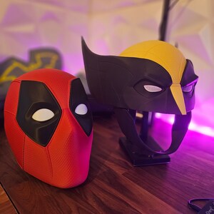 May include: Two superhero masks, one red and black with white eyes and a textured surface, and the other black and yellow with white eyes and a textured surface. The masks are on a wooden surface.