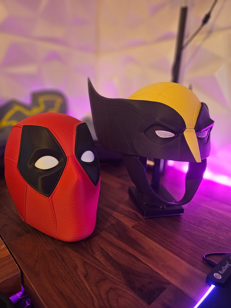May include: Two superhero masks, one red and black with white eyes and a textured surface, and the other black and yellow with white eyes and a textured surface. The masks are on a wooden surface.