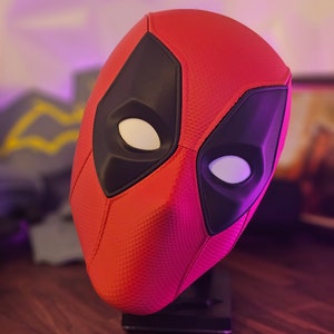 May include: A red and black Deadpool mask with a textured surface. The mask is on a black stand.