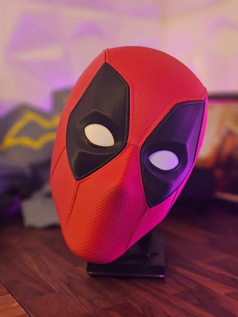 May include: A red and black Deadpool mask with a textured surface. The mask is on a black stand.