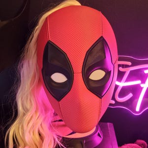 May include: A red and black Deadpool mask with a blonde wig attached to the back. The mask has a textured surface and a black eye mask with white eyes.