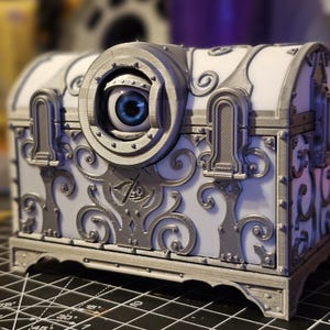 Peut inclure: Un coffre au trésor décoratif blanc et argent avec un design orné et un grand œil sur le devant. Le coffre est en plastique imprimé en 3D.