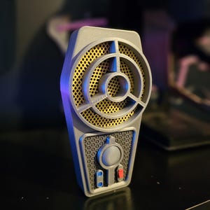 May include: A gray 3D printed handheld device with a yellow and black speaker grille, a round button, and two smaller blue buttons. The device has a textured surface and a red button.