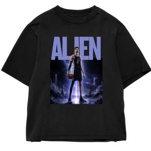 May include: Black t-shirt with the word "ALIEN" in light blue block letters. The shirt features a graphic of a basketball player holding a ball, set against a futuristic, otherworldly background with glowing lights and a misty atmosphere.