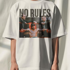 May include: White t-shirt featuring a graphic of a boxing scene with the text "NO RULES" in bold, dark grey letters. The image depicts a shirtless boxer in red gloves, surrounded by two men in suits. The shirt has short sleeves.
