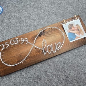 Könnte beinhalten: Ein Holzschild mit einem Unendlichkeitszeichen und dem Wort "love", das mit weißem Garn bespannt ist. Das Datum "25.03.99" ist ebenfalls auf dem Schild gespannt. Ein Foto eines Paares ist mit einer Wäscheklammer an dem Schild befestigt.