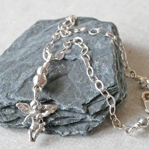 Puede incluir: Un brazalete de plata con un peque&ntilde;o dije de &aacute;ngel y cuentas de plata. El brazalete est&aacute; sobre un fondo de piedra gris.