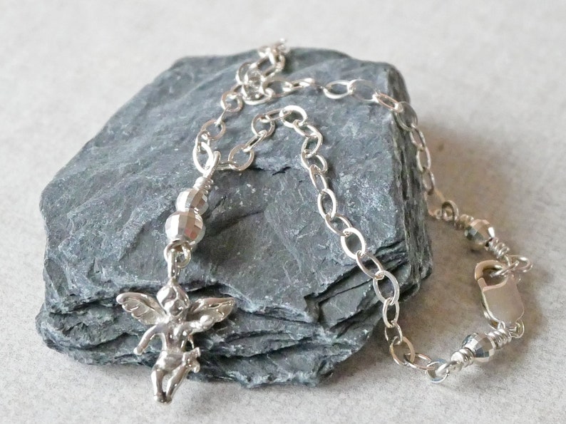 Puede incluir: Un brazalete de plata con un peque&ntilde;o dije de &aacute;ngel y cuentas de plata. El brazalete est&aacute; sobre un fondo de piedra gris.
